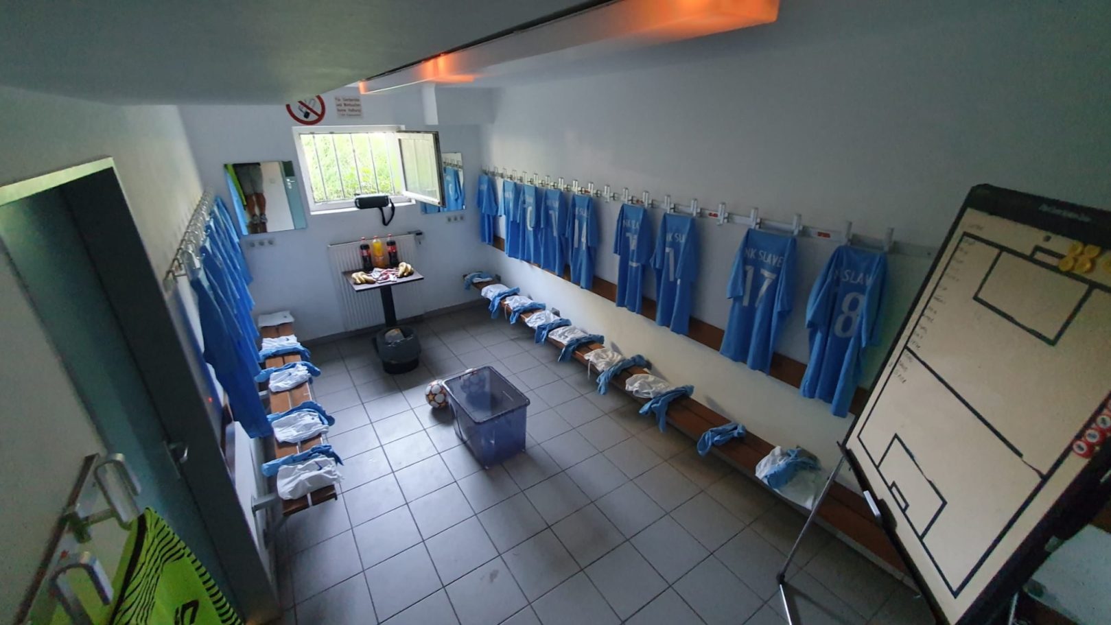 Umkleidekabine mit blauen Trikots, Bänken und einem Taktikboard an der Wand.