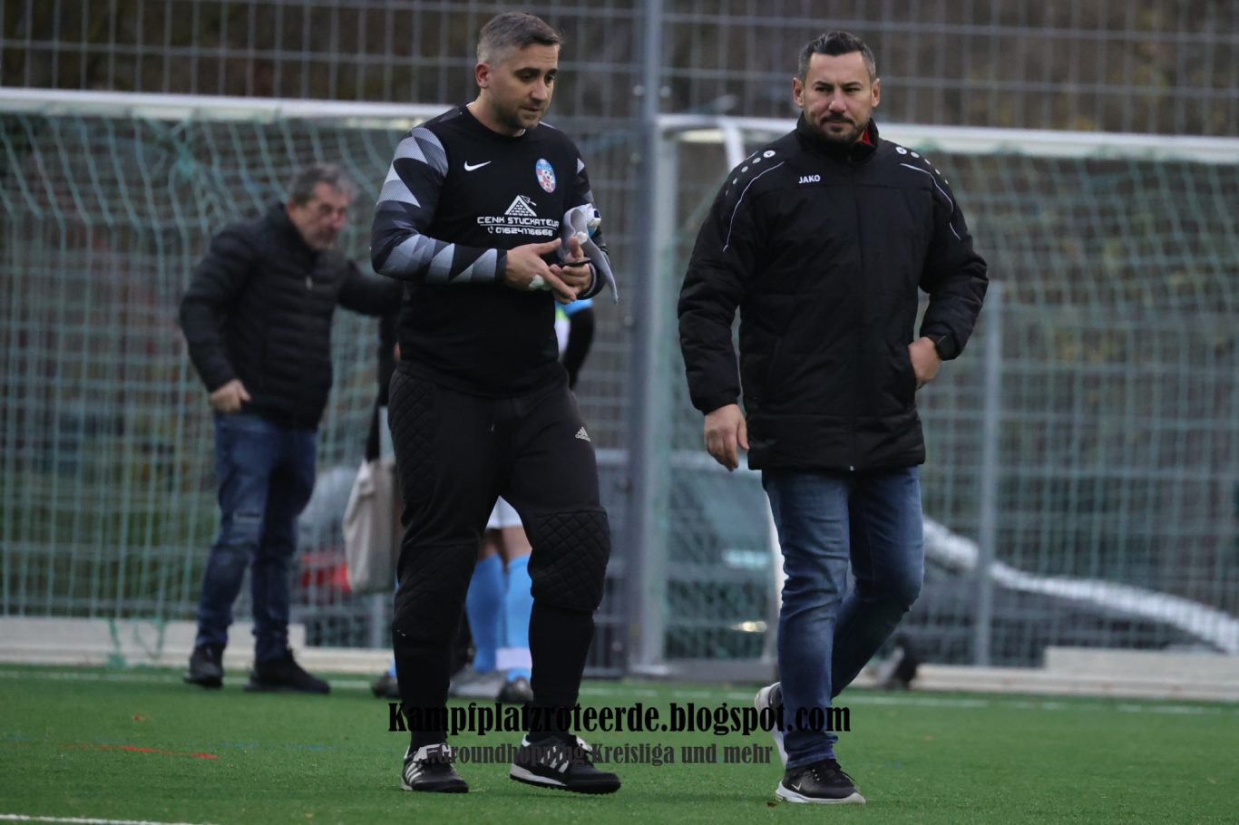 Zwei Männer in Sportkleidung gehen auf einem Fußballfeld, im Hintergrund weitere Personen.