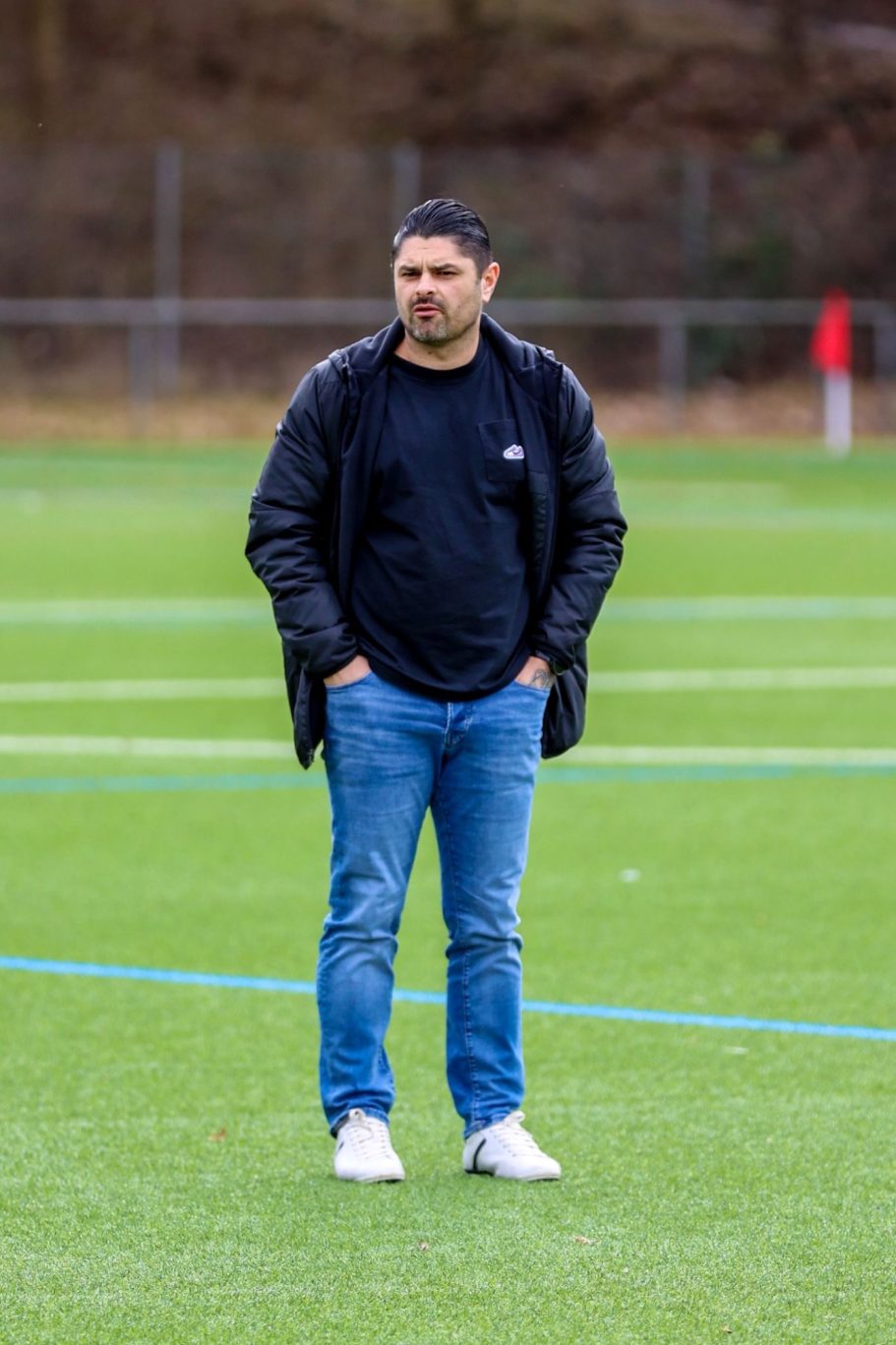 Mann in Jeans und Jacke steht auf einem Sportplatz mit grüner Rasenfläche.