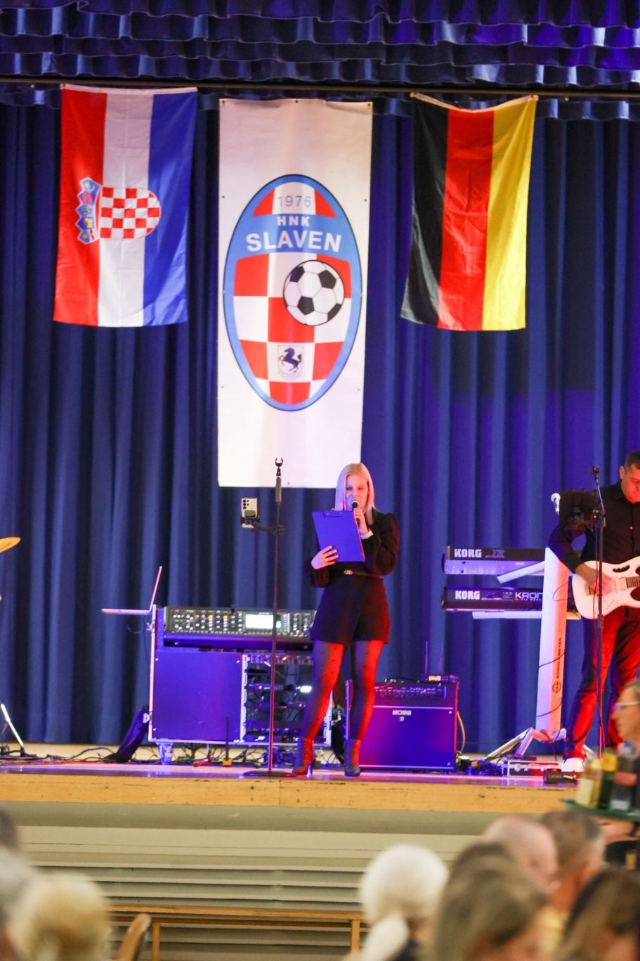 Frau mit Mikrofon auf der Bühne, im Hintergrund Flaggen von Kroatien und Deutschland.